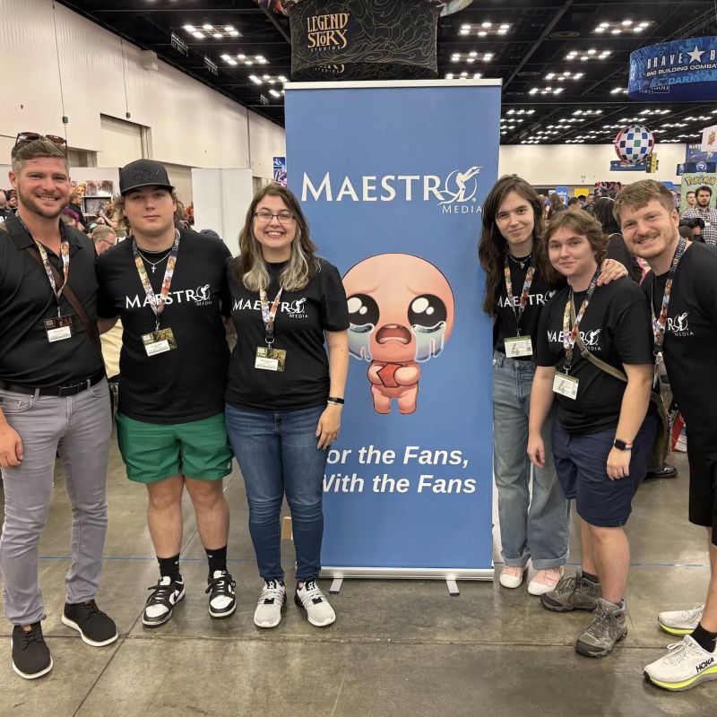 The Maestro Media team at Gen Con 2026 in front of a Binding of Isaac banner reading For the Fans, With the Fans